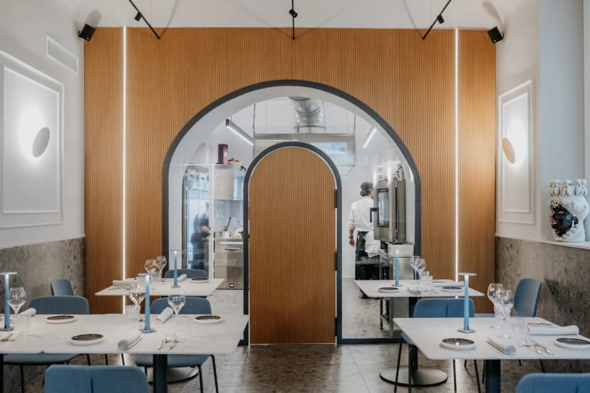 La sala di Dòmini: tavoli apparecchiati, pareti in legno e, in fondo, l'arco con la vetrata sulla cucina