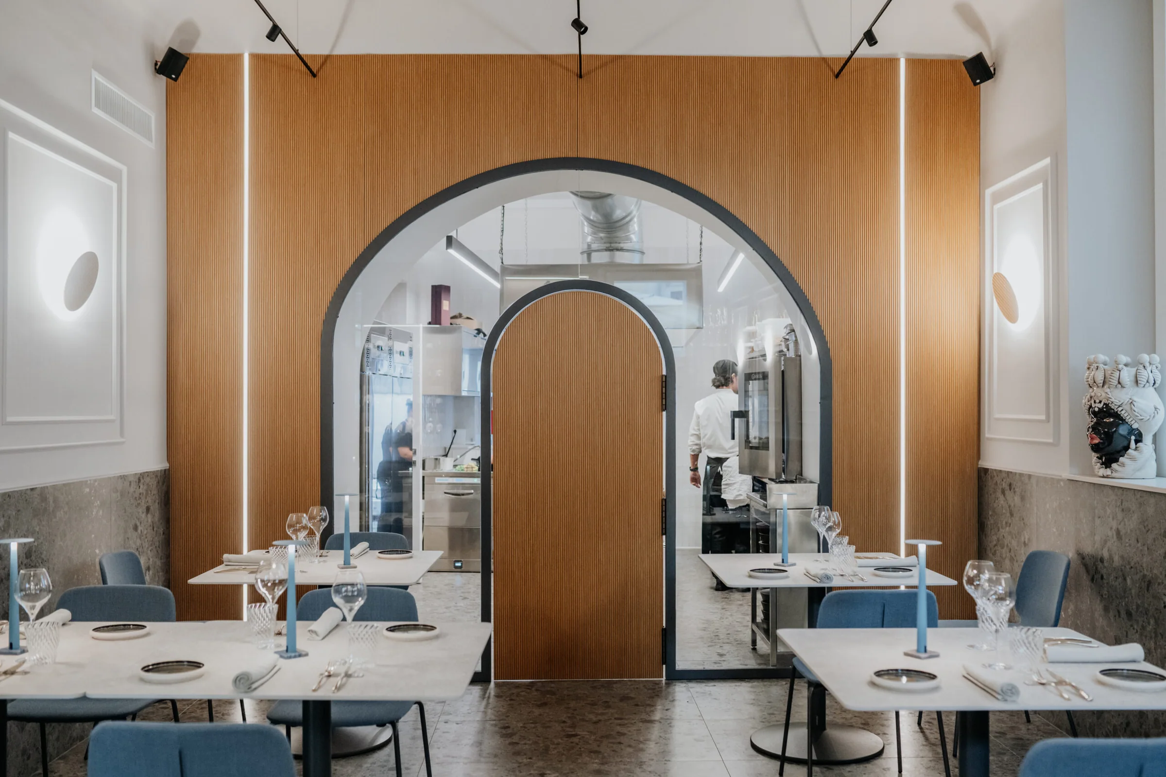 La sala di Dòmini: tavoli apparecchiati, pareti in legno e, in fondo, l'arco con la vetrata sulla cucina